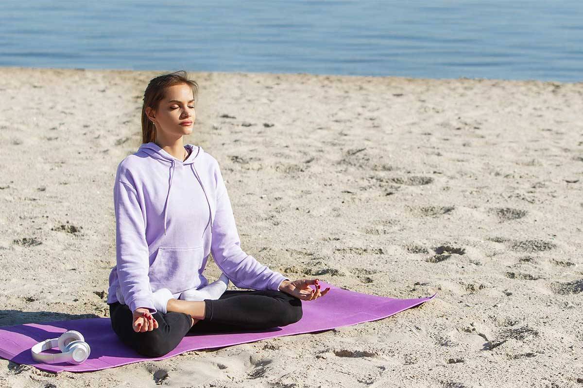 Eine junge Frau in violetten Sportklamotten sitzt am Strand und macht Yoga im Ostseeurlaub in den Arkona Strandresidenzen.