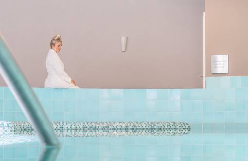Eine blonde Frau sitzt am Beckenrand des Hotelpools bereit für die Wassergymnastik im Ostseeurlaub in den Arkona Strandresidenzen.