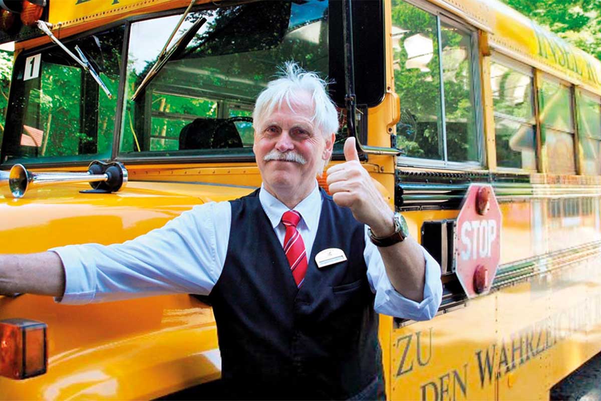 Ein Mann in Uniform lächelt in die Kamera und steht vor einem gelben Schulbus bereit für einen Ausflug der Arkona Strandresidenzen.